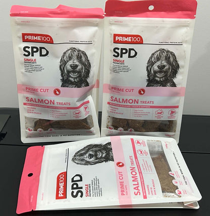 Three stand-up pouches of PRIME100 SPD Single and Large salmon treats for dogs, arranged on a dark surface against a light gray background.